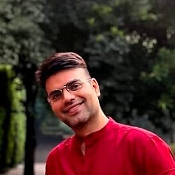 BK Sahni photographed outdoors, head and shoulders, in a red kurta against green foliage.